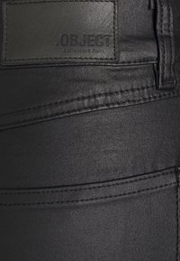 Jean en denim noir avec une patch en cuir portant le texte « .OBJECT collectors item », des coutures apparentes et une texture lisse.