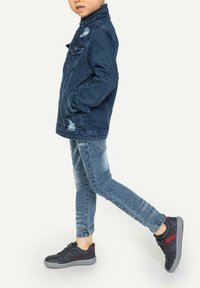 Veste en denim avec des détails usés, associée à un jean skinny bleu et des baskets noires avec des accents rouges. Coupe décontractée, matériaux résistants.