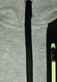 Grijze hoodie met rits en zwarte rits, voorzien van zwarte zijpanelen en een neon groene ritssluiting aan de rechterzak. Zachte textuur.