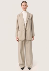 Beige szabott zakó, bemetszett hajtókával, két gombbal, oldalzsebekkel, laza szabású bézs nadrággal és fekete hegyes orrú magassarkú cipővel párosítva.