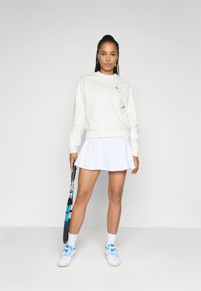 Sweat-shirt blanc à col rond et petit logo, associé à une jupe plissée blanche avec un design perforé. Tenant une raquette de tennis.