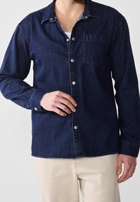 Mörk denimskjorta med spetskrage, metallknappar, bröstficka och långa ärmar. Slät textur och klassisk design.