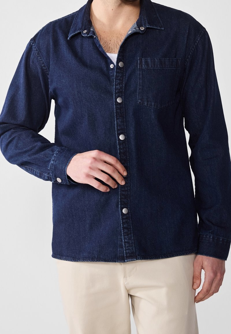 Mörk denimskjorta med spetskrage, metallknappar, bröstficka och långa ärmar. Slät textur och klassisk design.