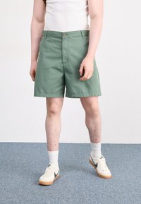 Pantalones cortos de algodón verde con un frente plano, dos bolsillos delanteros y cierre de botón, combinados con calcetines blancos y zapatillas beige.