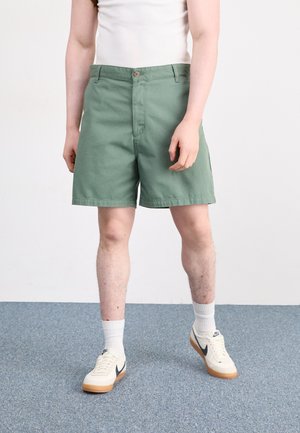Pantalones cortos de algodón verde con un frente plano, dos bolsillos delanteros y cierre de botón, combinados con calcetines blancos y zapatillas beige.