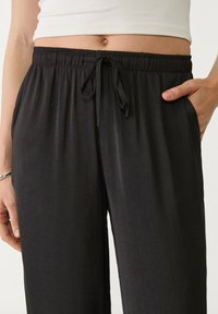 Žena v černých kalhotách s elastickým pasem a šňůrkou, s rukama v kapsách, v kombinaci s krčeným bílým topem, který odhaluje břicho.