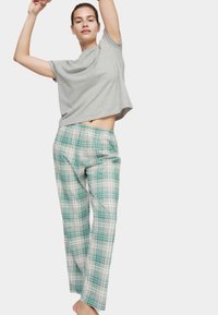 Mujer con camiseta gris estirando los brazos hacia arriba, vistiendo pantalones de pijama sueltos de cuadros verde y blanco contra un fondo sencillo.