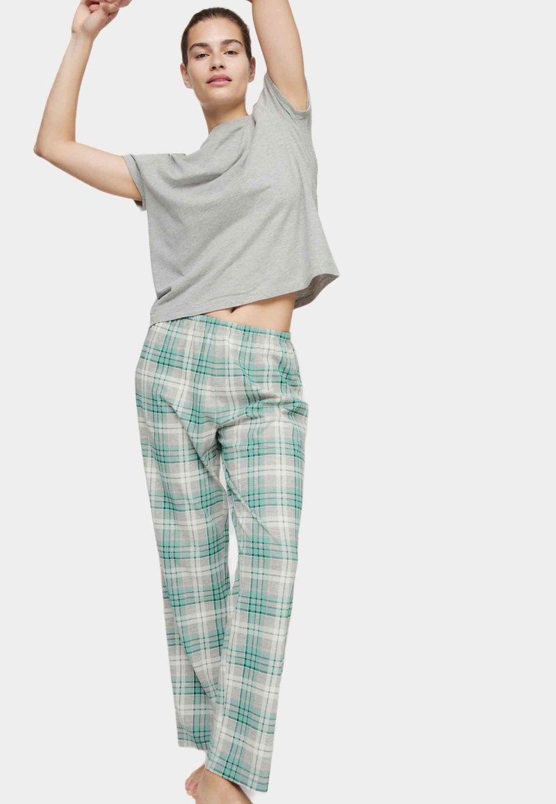 Mujer con camiseta gris estirando los brazos hacia arriba, vistiendo pantalones de pijama sueltos de cuadros verde y blanco contra un fondo sencillo.