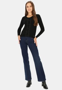 Czarny długi sweter dzianinowy z rękawami, z przodem na guziki, subtelną strukturą i dopasowanym krojem, w połączeniu z ciemnoniebieskimi dżinsami dzwonami i czarnymi botkami.