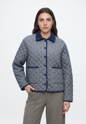 Femme portant une veste matelassée à carreaux bleue et grise avec col et bordure marine, associée à un pantalon gris à jambes larges.