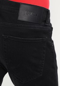 Černé džíny z denimu s koženou nášivkou na pásu s vyraženým názvem značky "EDWIN". Obsahují jednu zadní kapsu s ozdobným prošíváním.