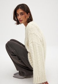 Knit cream sweater with textured pattern, oversized sleeves, paired with dark gray trousers and sleek gray shoes, against a plain backdrop.