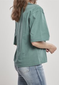 Tričko s krátkým rukávem, oversized, v tlumené zelené barvě z bavlny, s kulatým výstřihem a hladkou texturou. Nosené s světlými modrými džíny.