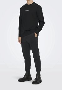 Camicetta nera a maniche lunghe con un piccolo logo bianco, abbinata a pantaloni neri affusolati e stivaletti neri, con polsini elasticizzati e tessuto testurizzato.