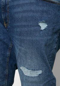 Jean en denim bleu foncé avec des patchs usés. Des trous effilochés notables dans une teinte de bleu plus clair, présentant des variations de texture dues à l'usure.