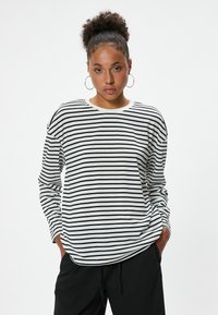 Gestreiftes Langarmshirt in Schwarz und Weiß mit rundem Halsausschnitt; lockere Passform; der Stoff wirkt weich mit leichtem Glanz; getragen mit schwarzen Hosen.