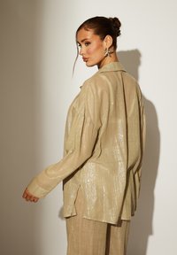 Camisa beige, texturizada y transparente con un acabado brillante, que presenta aberturas laterales y puños abotonados. Llevada sobre una capa base a juego.