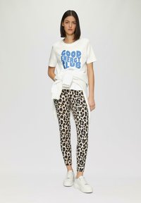 Witte t-shirt met blauwe "GOOD ENERGY CLUB" graphic, gecombineerd met legging met luipaardprint. Model draagt witte platform sneakers. Simpe achtergrond.