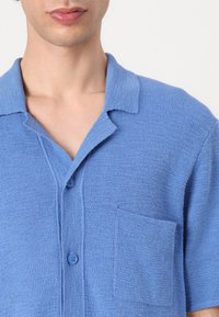 Cămașă din bumbac albastră cu mânecă scurtă, dintr-un material texturat, cu guler deschis și două butoane. Dispune de un buzunar aplicat pe partea stângă.