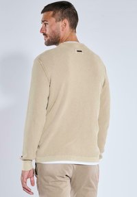 Beige gerippter Pullover mit langen Ärmeln, Rundhals und dezentem Logo-Detail auf der Rückseite, kombiniert mit hellen Hosen.