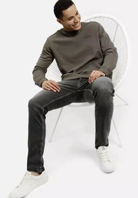 Mężczyzna uśmiechający się, siedzący na białym nowoczesnym krześle, ubrany w szary sweter, ciemne dżinsy i białe sportowe buty na tle gładkiej białej ściany.