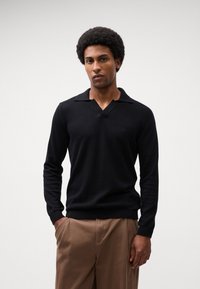 Zwarte, lange mouw pullover met een V-hals en ruime kraag. Zachte textuur, aansluitend ontwerp, gecombineerd met tan broeken. Subtiele logo-detail op de borst.