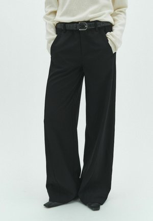 Persona con pantalones negros de pierna ancha, cinturón negro y jersey crema, con las manos en los bolsillos, de pie frente a un fondo liso.