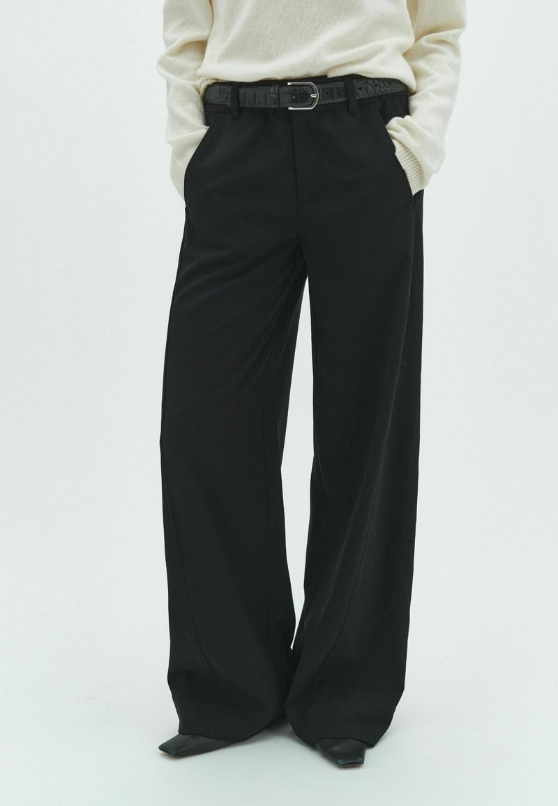 Personne portant un pantalon noir à jambes larges avec une ceinture noire et un pull crème, mains dans les poches, debout devant un fond uni.