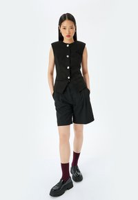 Zwarte mouwloze vest met een gestructureerd ontwerp, voorzien van zilveren knopen. Gecombineerd met zwarte shorts en platformschoenen met bordeauxrode sokken.
