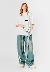 Camiseta blanca extragrande con estampado negro "MWM", combinada con vaqueros holgados azul claro con texto blanco y una riñonera gris con logotipos.