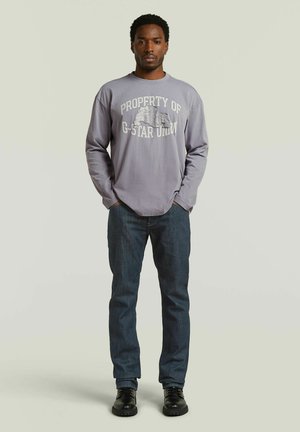 Man die staat met zijn handen in zijn zakken, draagt een grijs shirt met lange mouwen met de tekst "PROPERTY OF G-STAR UNION," donkere spijkerbroek en zwarte schoenen.