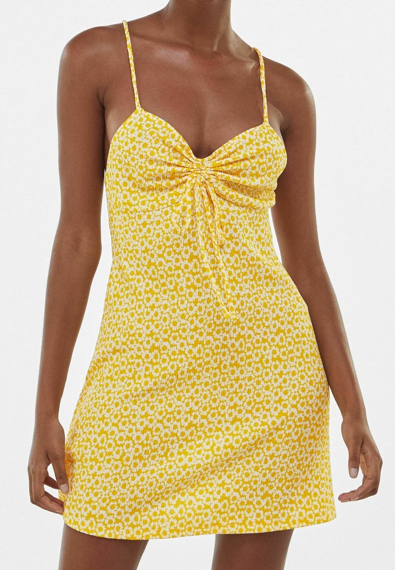 Femme portant une mini-robe jaune avec un imprimé marguerite blanche, fines bretelles et détail froncé au centre de la poitrine.