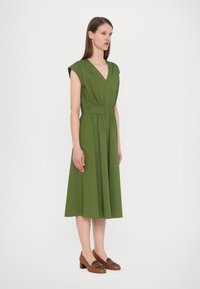 Vihreä polvipituinen mekko, jossa on V-kaula-aukko, lyhyet hihat ja selkeä vyötärö. Yhdistetty tummempiin ruskeisiin loafer-kenkiin.