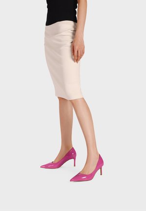 Mujer con falda lápiz beige y zapatos de tacón alto de charol rosa brillante, de pie sobre un fondo claro y liso.