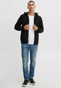 Schwarze Zip-Hoodie mit Kapuze und Kordelzug, getragen über einem weißen T-Shirt, kombiniert mit hellblauen Jeans und schwarzen Sneakers. Einfaches Design.