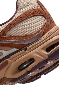 Primer plano de la suela y lateral de un zapato Nike Air Max marrón y beige, con tela de malla, superposiciones de ante y unidad de amortiguación de aire visible.