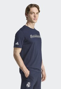 Camiseta de algodón azul marino con mangas cortas, que presenta el texto "Real Madrid" en blanco y un logo de Adidas en la manga izquierda. Textura suave.