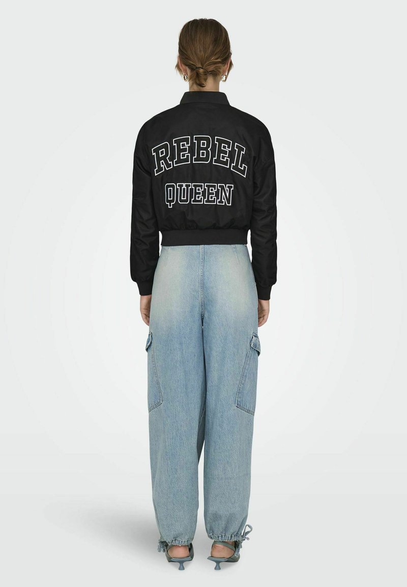 Veste bomber noire avec "REBEL QUEEN" en grandes lettres blanches au dos, associée à un jean large bleu clair et des sandales à talons vertes.