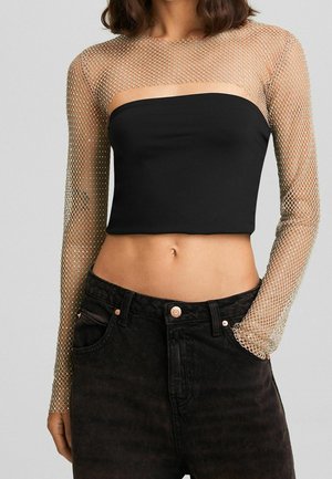 Femme portant un crop top noir sans bretelles superposé à une chemise en mesh transparente à manches longues et un jean noir taille haute, main dans la poche.