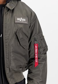 Olive green bomber jacket with smooth nylon fabric, featuring a front patch, zippered pockets, and a red "Remove Before Flight" tag.