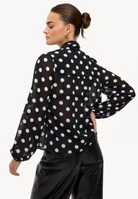Blouse noire transparente à gros pois blancs, manches longues bouffantes et col plissé. Portée avec un pantalon noir en similicuir brillant.