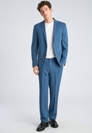 Jonge man die staat, gekleed in een blauw pak, wit t-shirt en zwarte nette schoenen, tegen een effen lichte achtergrond.