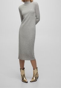 Robe en tricot côtelé grise avec un col montant et des manches longues, se terminant au milieu du mollet. Portée avec des bottines dorées brillantes à bout pointu.