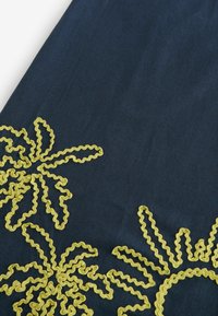 Tissu bleu marine avec broderie jaune ondulée formant des motifs floraux abstraits le long des bords.