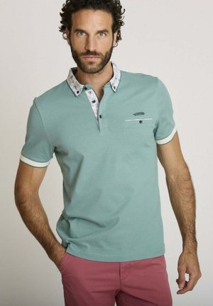 Homme aux cheveux bouclés et foncés portant un polo vert menthe avec un liseré blanc et un pantalon rose, debout avec une main dans la poche.
