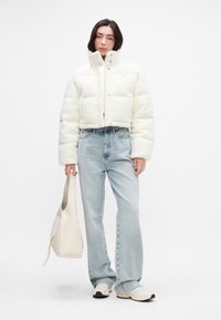 Ostrihaná biela fleecová puffer bunda s vysokým golierom a cvokmi, v kombinácii s svetlomodrými džínami s vysokým pásom a krémovou kabelkou.