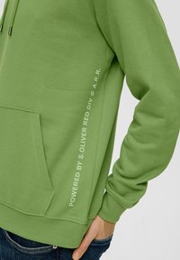 Sudadera verde con bolsillo canguro, puños acanalados y texto impreso en el lateral. Tejido suave y texturizado, corte relajado.