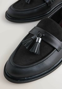 Czarne loafersy z zamszowej cholewki, ozdobione skórzanym paskiem oraz dwoma czarnymi frędzlami i metalowym akcentem. Okrągły czubek i teksturowana podeszwa.