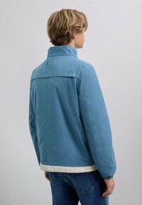Chaqueta azul claro con cuello alto, puños elásticos y un ribete crema en la cintura. Textura suave con un sutil detalle de logo en la parte trasera.