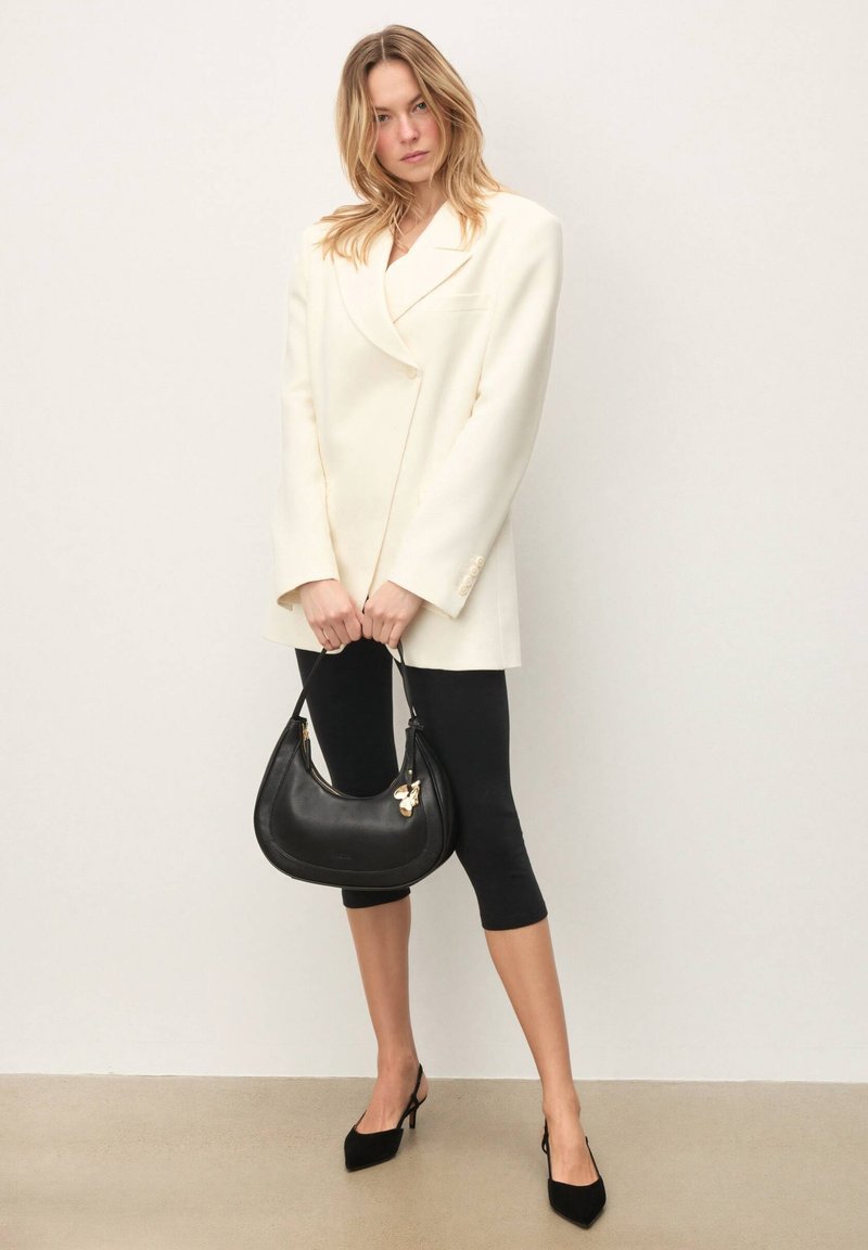 Veste oversize crème, leggings noirs et escarpins noirs à bout pointu. Tient un sac en cuir noir avec un accent doré. Textures lisses partout.